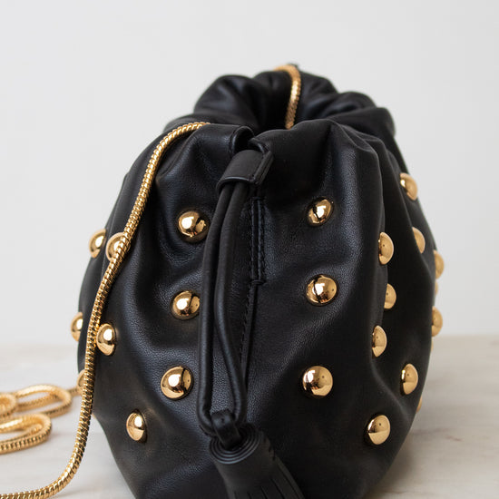 A black leather drawstring bag adorned with gold-tone studs and a gold chain strap, resting on a light marble surface against a white background.