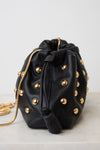 A black leather drawstring bag adorned with gold-tone studs and a gold chain strap, resting on a light marble surface against a white background.