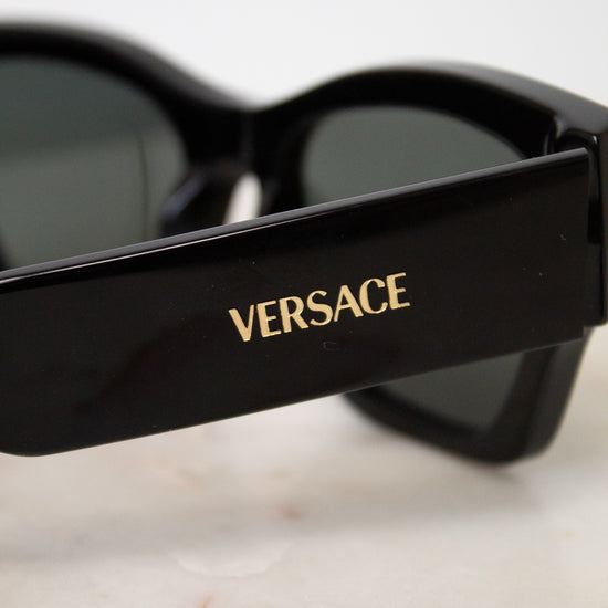 Close-up of black Versace sunglasses with dark lenses and gold logo lettering on the temple, resting on a light marble surface.