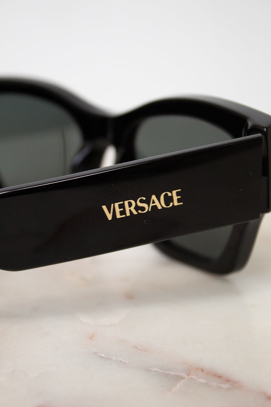 Close-up of black Versace sunglasses with dark lenses and gold logo lettering on the temple, resting on a light marble surface.