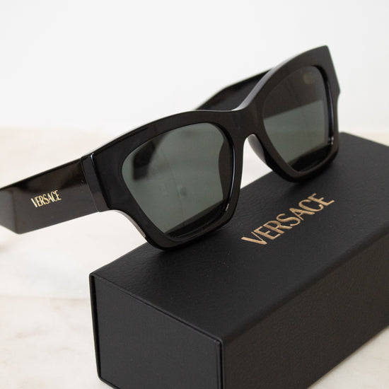 Black Versace sunglasses with dark lenses resting on top of a black Versace-branded box, set against a clean white background.
