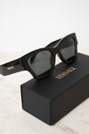 Black Versace sunglasses with dark lenses resting on top of a black Versace-branded box, set against a clean white background.