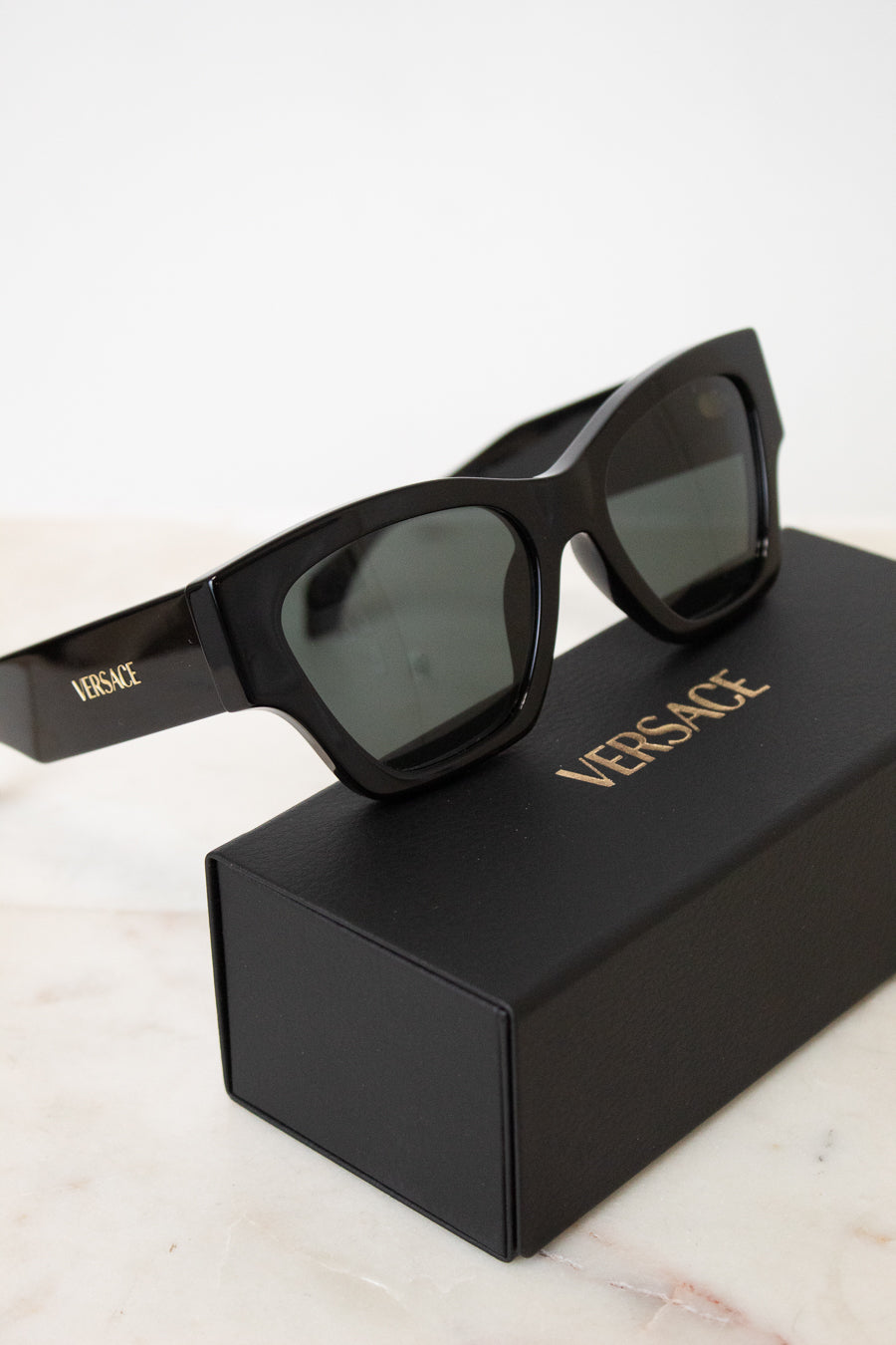 Black Versace sunglasses with dark lenses resting on top of a black Versace-branded box, set against a clean white background.