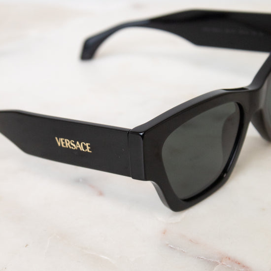 Black Versace sunglasses with dark lenses and gold logo lettering, resting on a white marble surface.