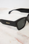 Black Versace sunglasses with dark lenses and gold logo lettering, resting on a white marble surface.