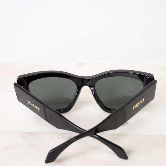 Black cat-eye sunglasses with dark lenses and gold VERSACE branding on the temples, resting on a light marble surface against a white background.