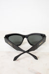 Black cat-eye sunglasses with dark lenses and gold VERSACE branding on the temples, resting on a light marble surface against a white background.