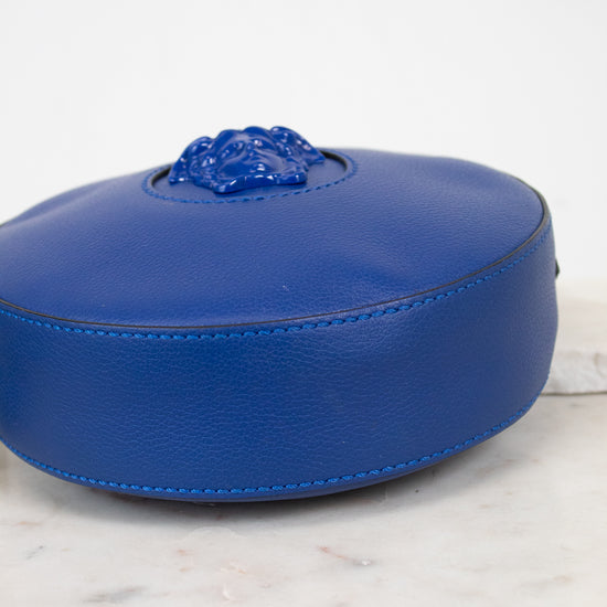 A vibrant blue leather round bag with a sculpted medusa head detail, viewed from the bottom and resting on a white marble surface against a white background.