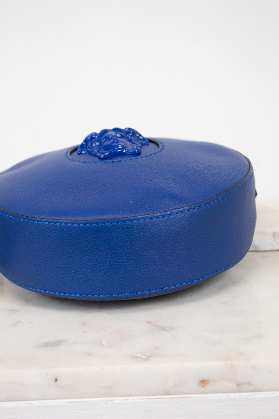 A vibrant blue leather round bag with a sculpted medusa head detail, viewed from the bottom and resting on a white marble surface against a white background.