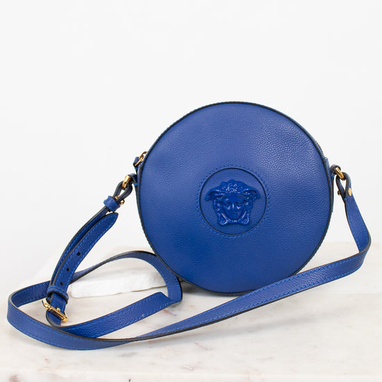 A blue leather crossbody bag with a circular body, attached blue Medusa logo on the front, and a matching blue strap, displayed on a white marble surface against a white background.
