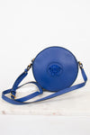 A blue leather crossbody bag with a circular body, attached blue Medusa logo on the front, and a matching blue strap, displayed on a white marble surface against a white background.