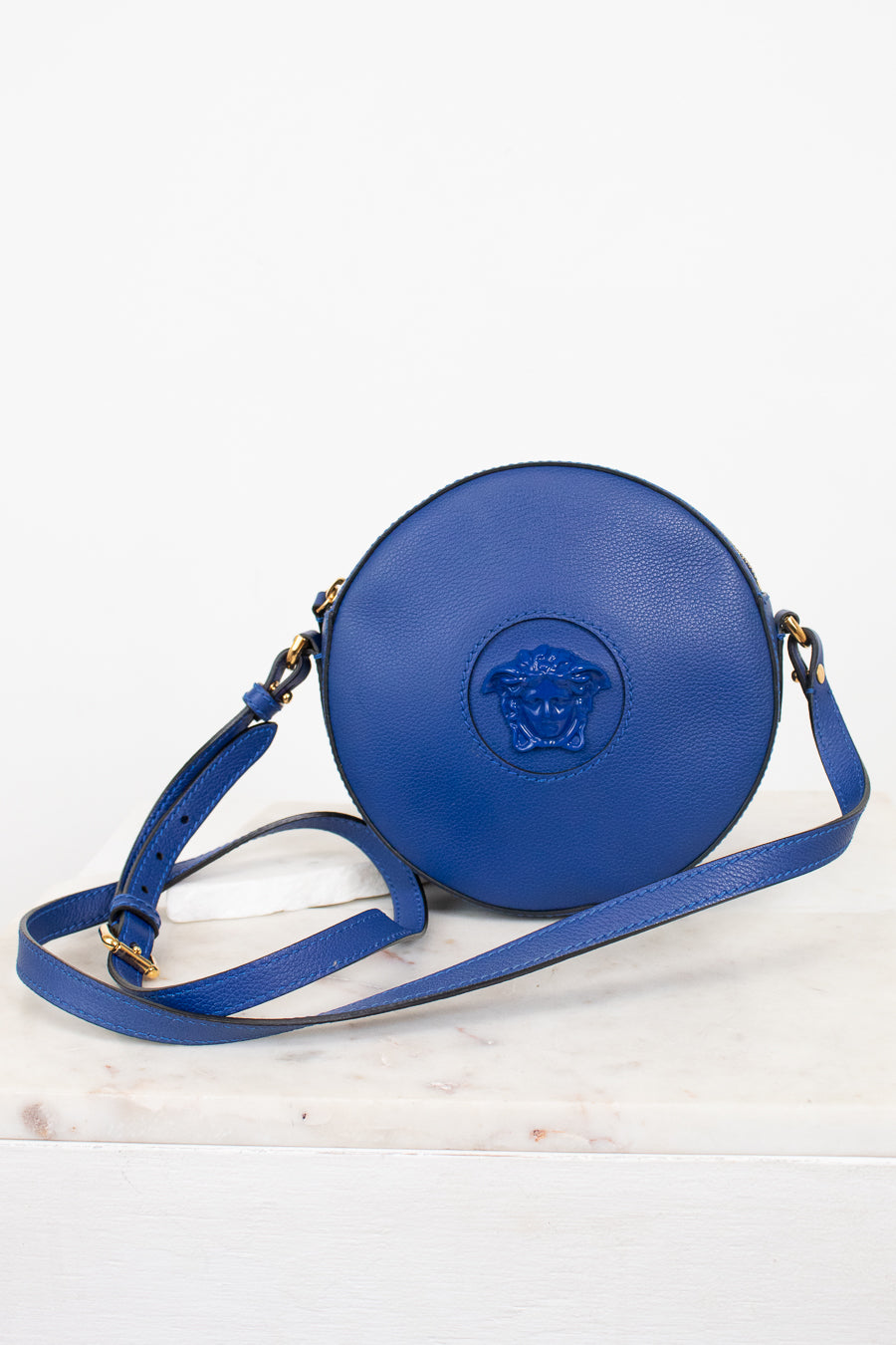 A blue leather crossbody bag with a circular body, attached blue Medusa logo on the front, and a matching blue strap, displayed on a white marble surface against a white background.