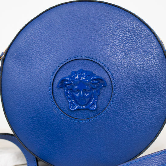 A round blue leather crossbody bag featuring a blue attached Medusa head logo on the front, with a matching blue strap laid across the lower portion of the frame against a light background.