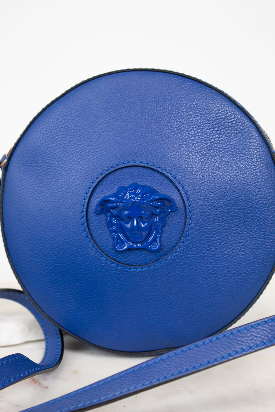 A round blue leather crossbody bag featuring a blue attached Medusa head logo on the front, with a matching blue strap laid across the lower portion of the frame against a light background.