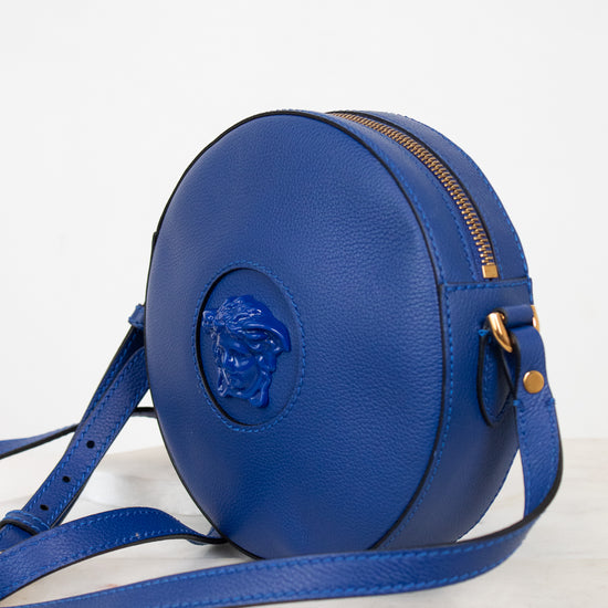 A round blue leather crossbody bag with a gold zipper and a prominent blue Medusa logo on the front, displayed on a light marble surface against a white background.