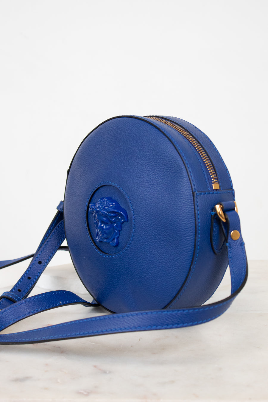 A round blue leather crossbody bag with a gold zipper and a prominent blue Medusa logo on the front, displayed on a light marble surface against a white background.