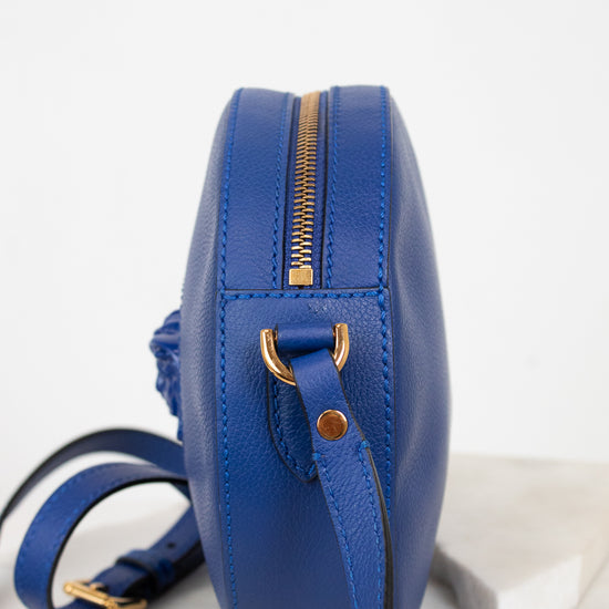 A royal blue leather crossbody bag with gold-tone hardware, shown from a side profile view on a light marble surface against a white background.