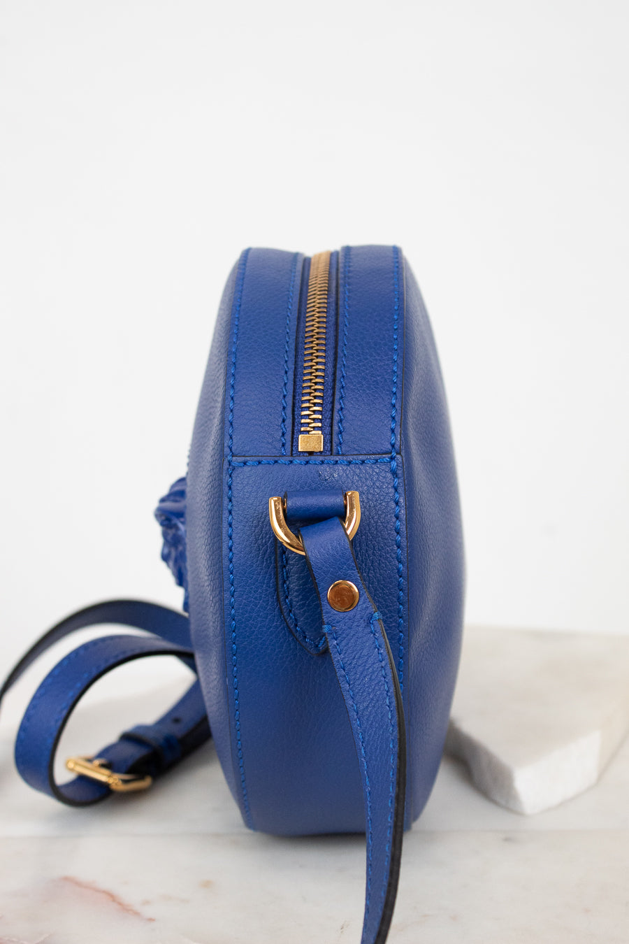 A royal blue leather crossbody bag with gold-tone hardware, shown from a side profile view on a light marble surface against a white background.