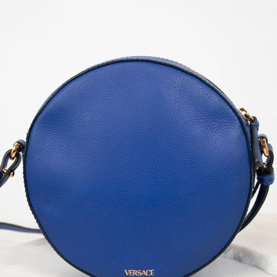 A round blue leather crossbody bag with gold-tone hardware and a small gold VERSACE logo on the back, displayed on a light marble surface against a white background.