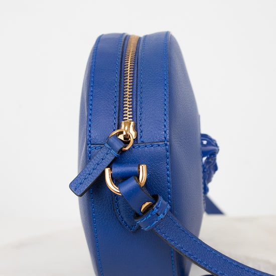 A small round blue leather crossbody bag with gold-tone hardware and a matching blue strap, shown from a side profile on a light marble surface against a white background.