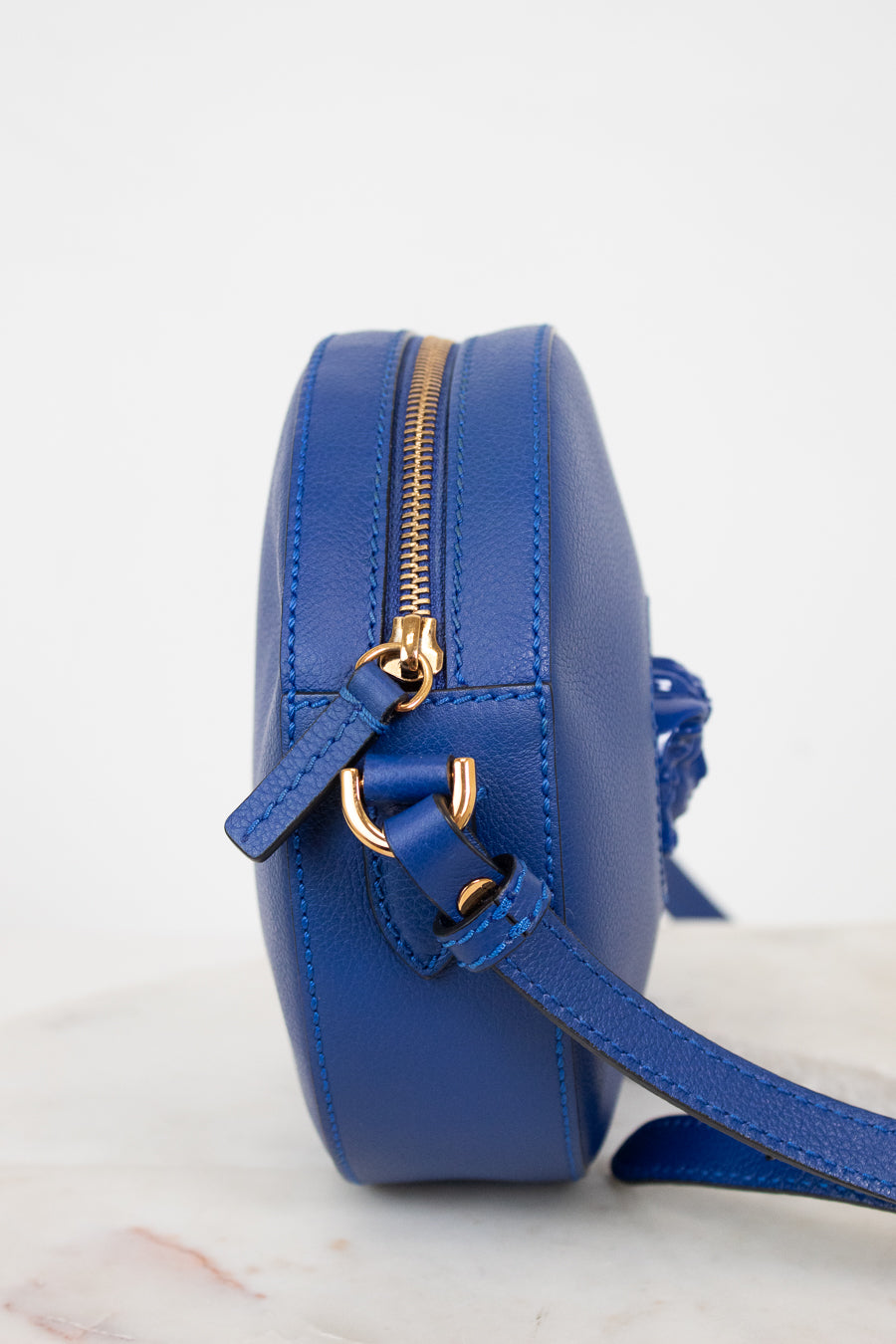 A small round blue leather crossbody bag with gold-tone hardware and a matching blue strap, shown from a side profile on a light marble surface against a white background.