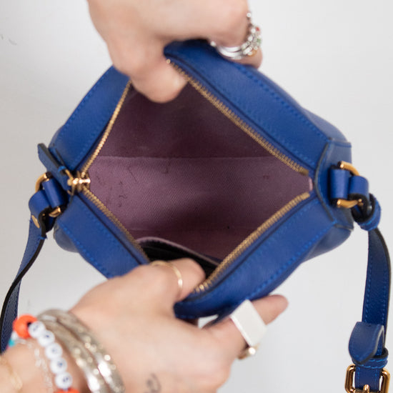 A close-up of a blue leather handbag being opened by two hands, showing the interior lining and gold-tone zipper hardware against a plain light background.