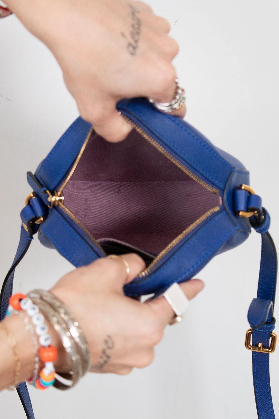 A close-up of a blue leather handbag being opened by two hands, showing the interior lining and gold-tone zipper hardware against a plain light background.