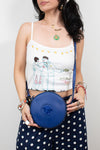 A woman wearing a white spaghetti-strap crop top with a graphic print and dark navy polka-dot pants, holding a round blue crossbody bag with a prominent blue Medusa logo. She is accessorized with gold necklaces, bracelets, and rings against a plain light gray background.