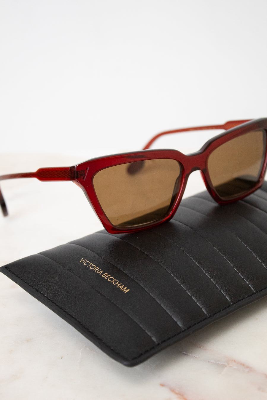 Victoria Beckham sunglasses with a translucent red acetate frame and brown tinted lenses, resting on a black quilted Victoria Beckham glasses case over a white marble surface.