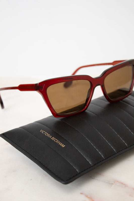 Victoria Beckham sunglasses with a translucent red acetate frame and brown tinted lenses, resting on a black quilted Victoria Beckham glasses case over a white marble surface.