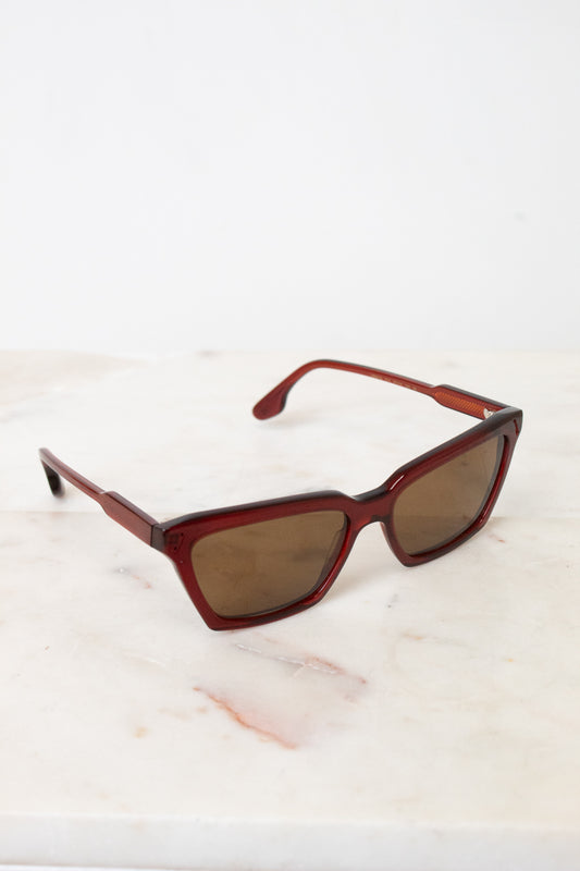 Retro-style cat-eye sunglasses with a translucent dark red acetate frame and brown tinted lenses, resting on a white marble surface against a white background.