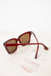 A pair of angular cat-eye sunglasses with a translucent dark red acetate frame and brown tinted lenses, resting on a light marble surface against a white background.