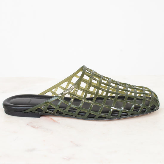 An olive green open-weave mule sandal with a black sole, displayed on a light marble surface against a white background.