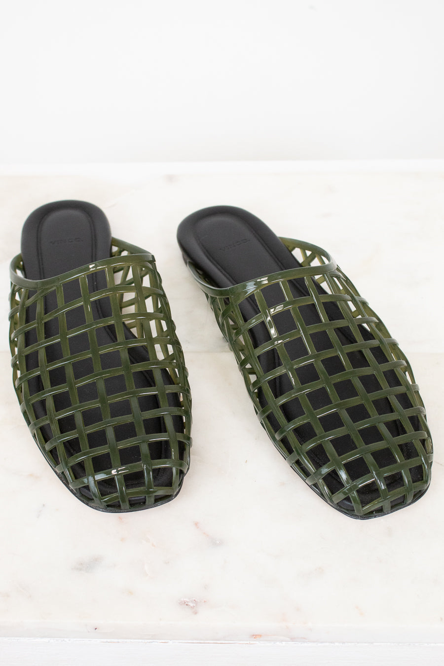 A pair of olive green open-toe mule sandals with a glossy grid pattern, displayed on a white marble surface against a white background.