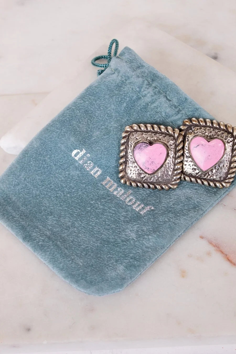 Pair of earrings with pink heart centers on a blue velvet pouch with 'Dian Malouf' branding.