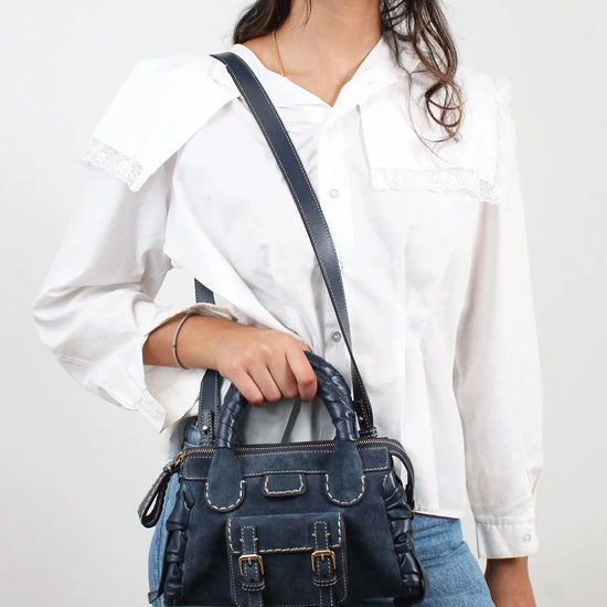 Person wearing a white shirt and blue jeans with a dark blue handbag on a plain background
