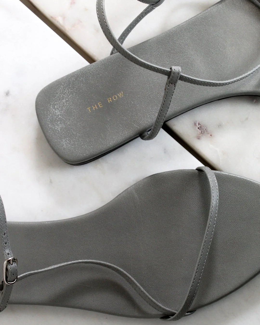 Gray sandals with 'The Row' branding on a marble surface