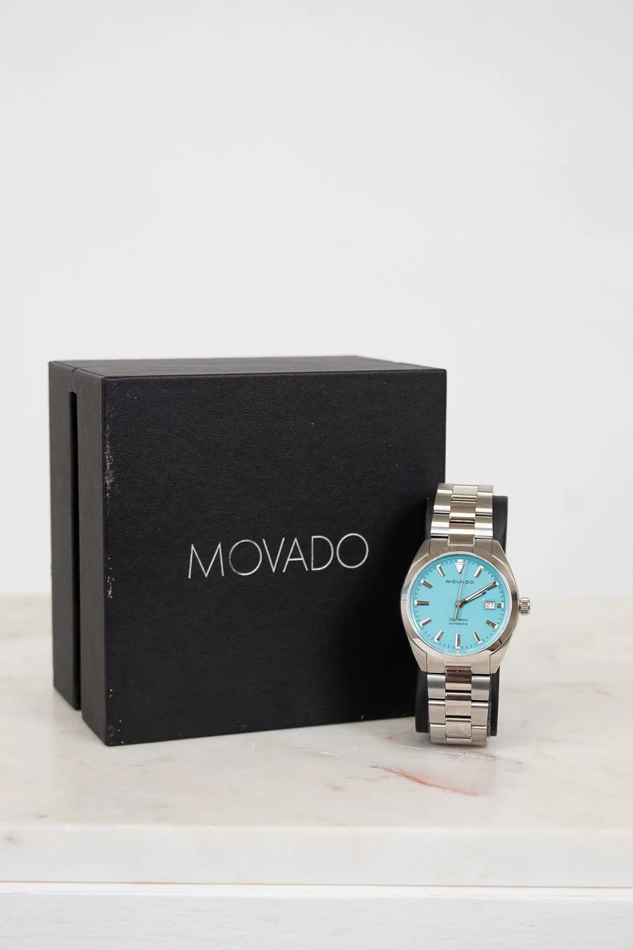 Movado watch with blue face and silver case next to a black Movado box on a white background