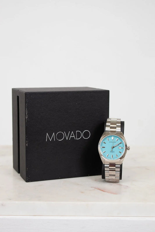 Movado watch with blue face and silver case next to a black Movado box on a white background