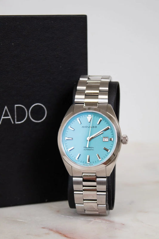 Silver watch with a blue face on a marble surface, brand box in the background