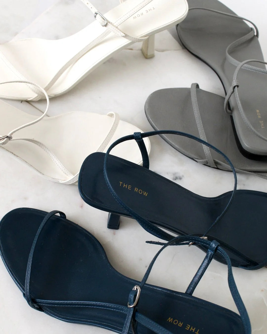 Navy, white, and gret  sandals with 'The Row' branding on a light marble background