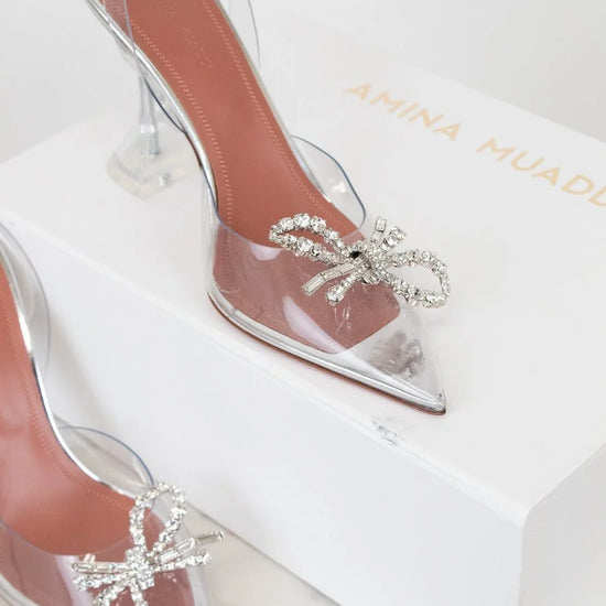 Clear high-heeled shoes with decorative bows on a branded box